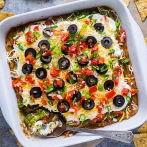 seven layer dip in square white dish with serving spoon and scattered tortilla chips