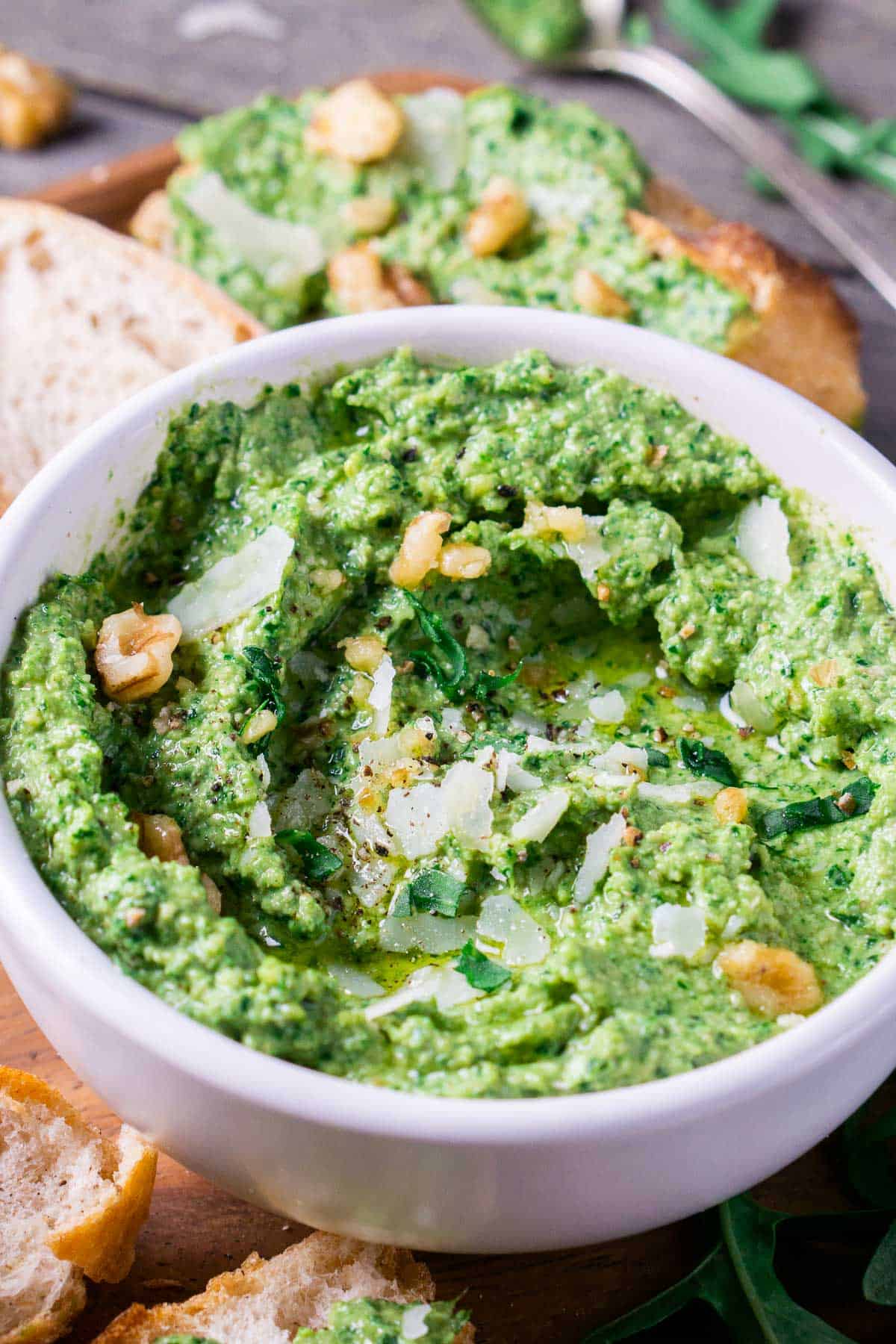 arugula pesto in white bowl on brown wood serving platter with sliced french bread