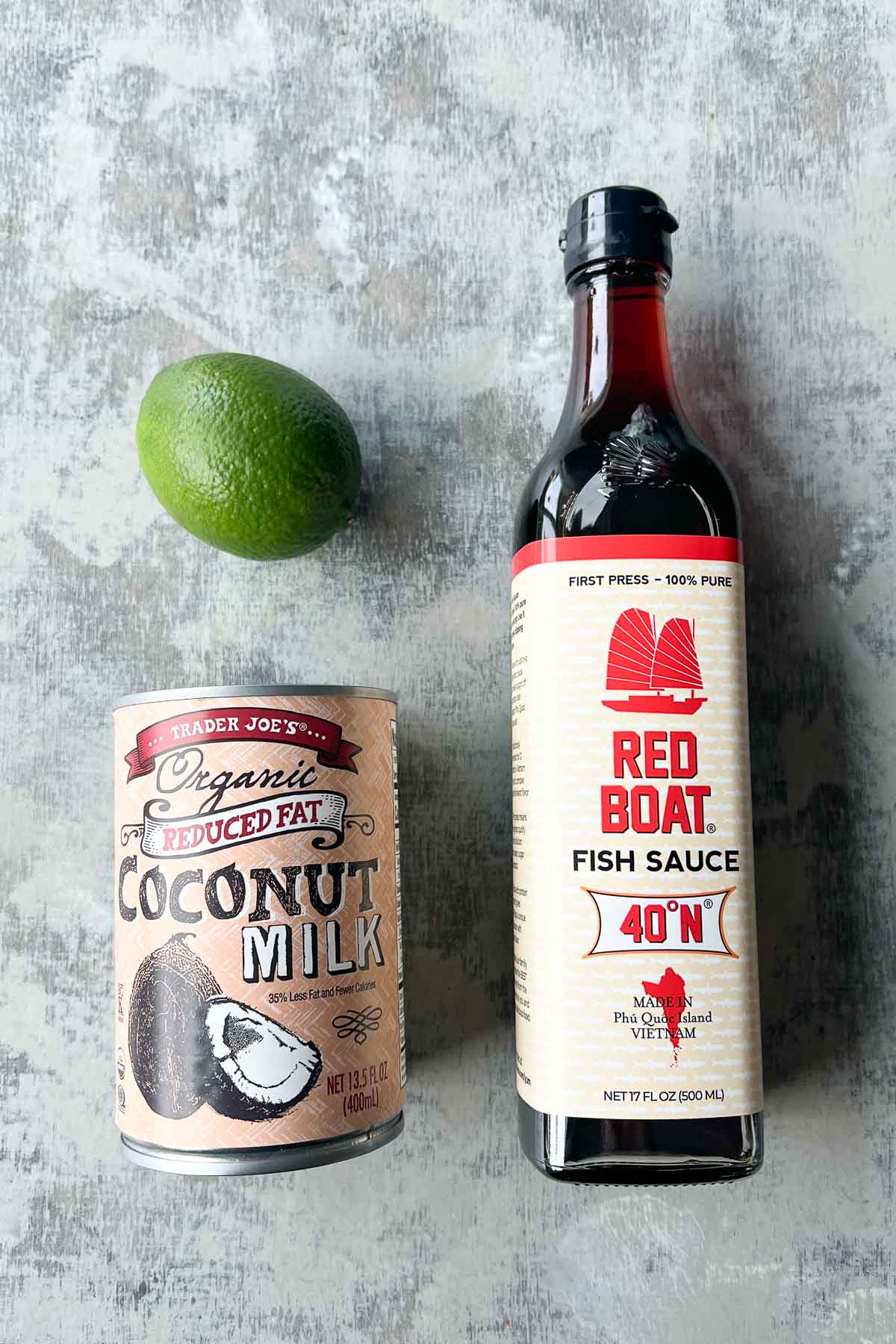 A lime, a can of coconut milk, and a bottle of fish sauce are arranged on a gray textured surface.