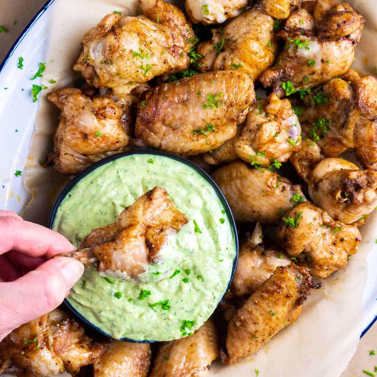 hand dunking chicken wing into bowl of avocado ranch dressing nested on platter of baked chicken wings