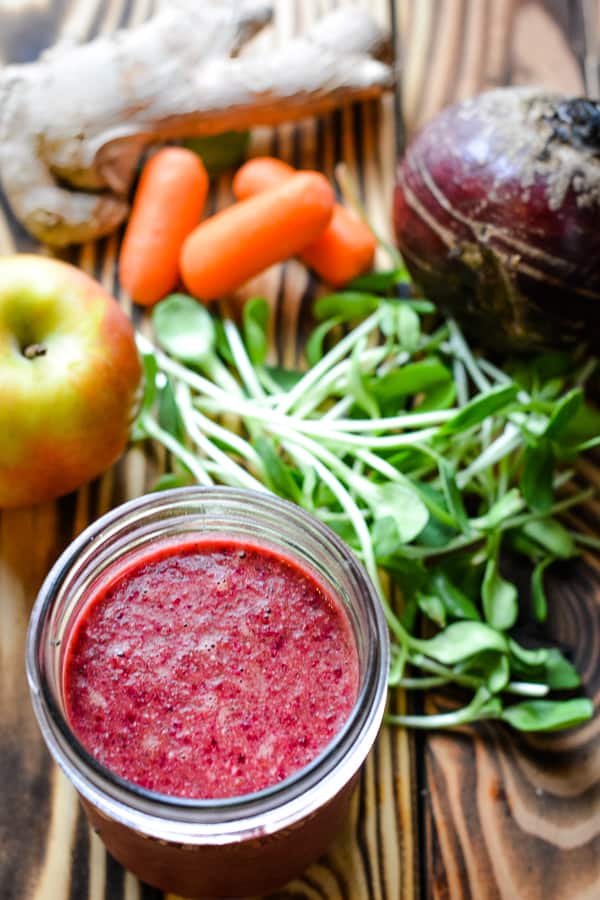 beet smoothie in glass jar with microgreens, whole apple, whole ginger, baby carrots, and whole beet on wood background