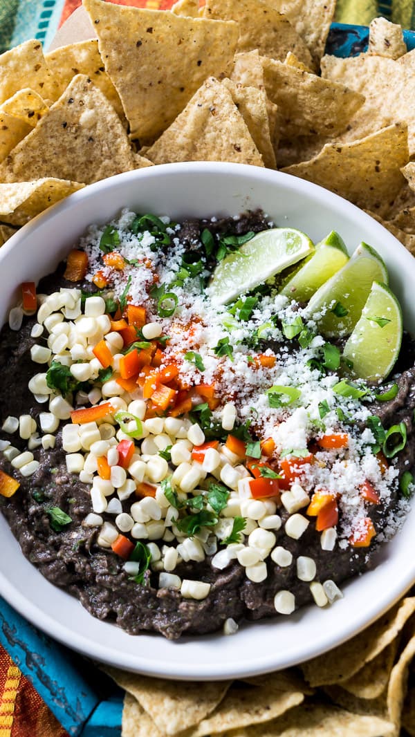 loaded black bean dip in white bowl surrounded by tortilla chips and multicolored background