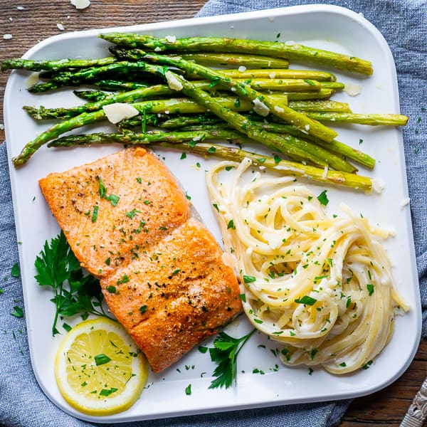 broiled salmon, sautéed asparagus, and fettuccini alfredo on white plate with lemon and parsley garnish