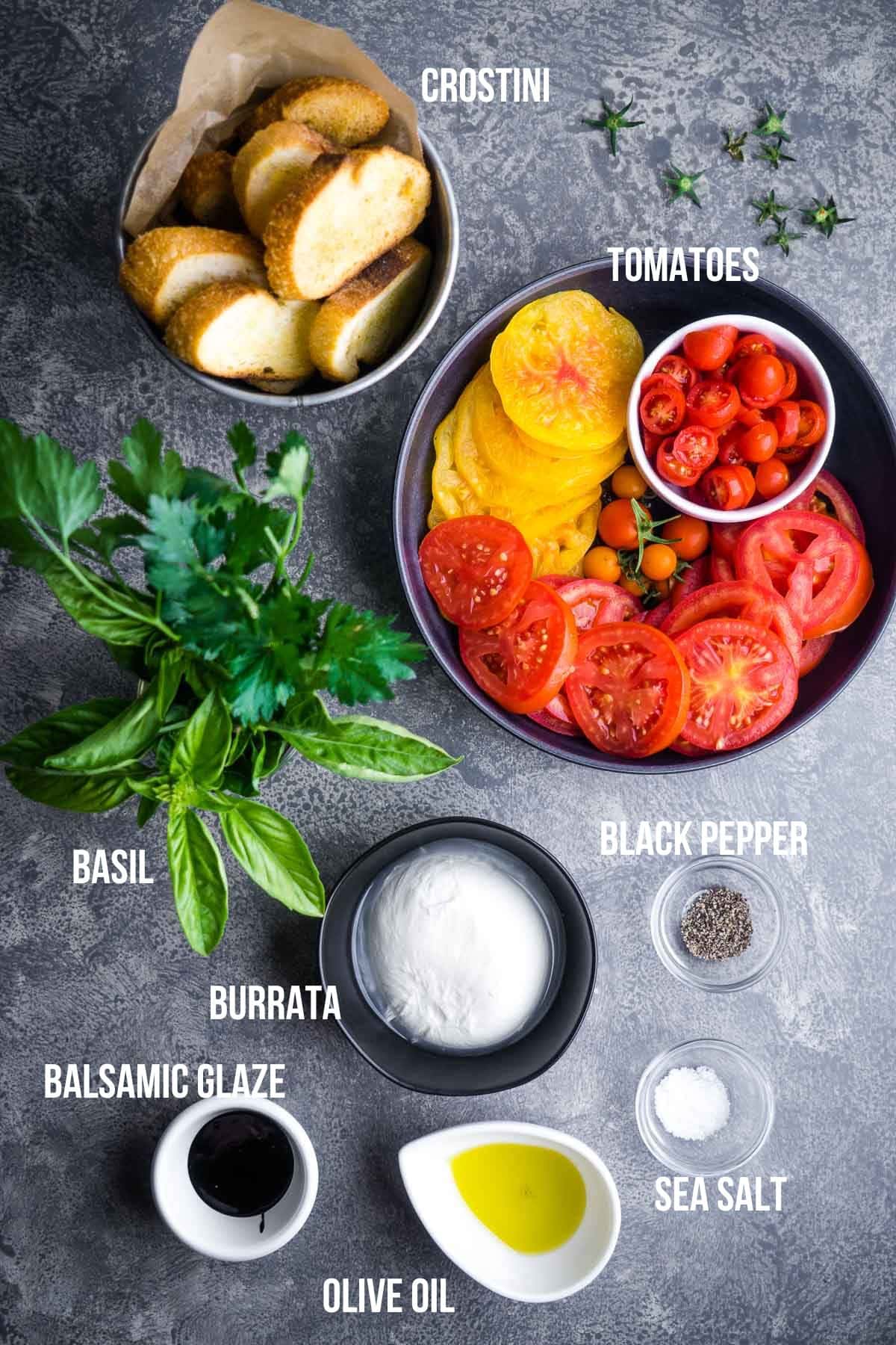 Overhead view of crostini, sliced tomatoes, cherry tomatoes, burrata, olive oil, balsamic glaze, sea salt, black pepper, and basil—each ingredient labeled for a vibrant tomato burrata salad.