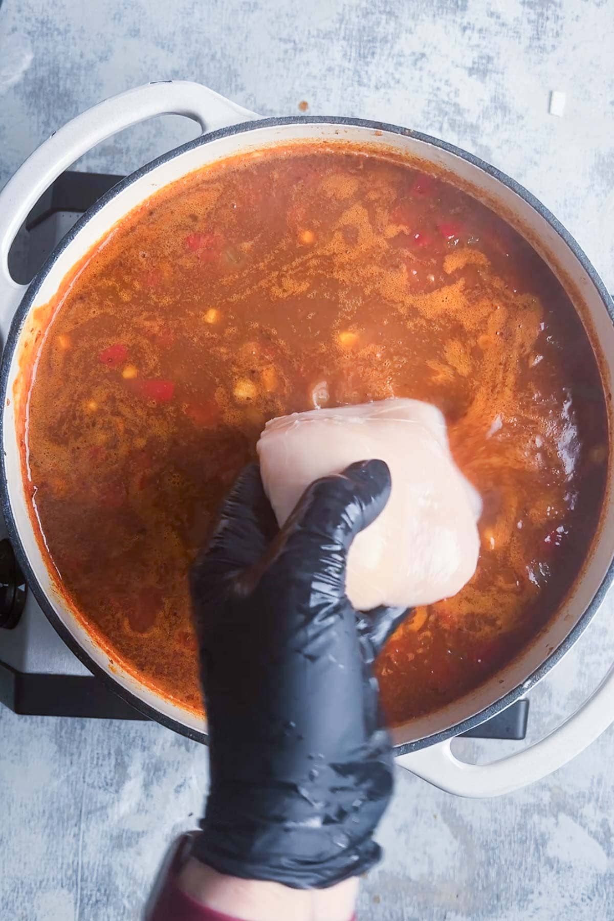 Gloved hand holding raw chicken over a pot of red soup on a stovetop.