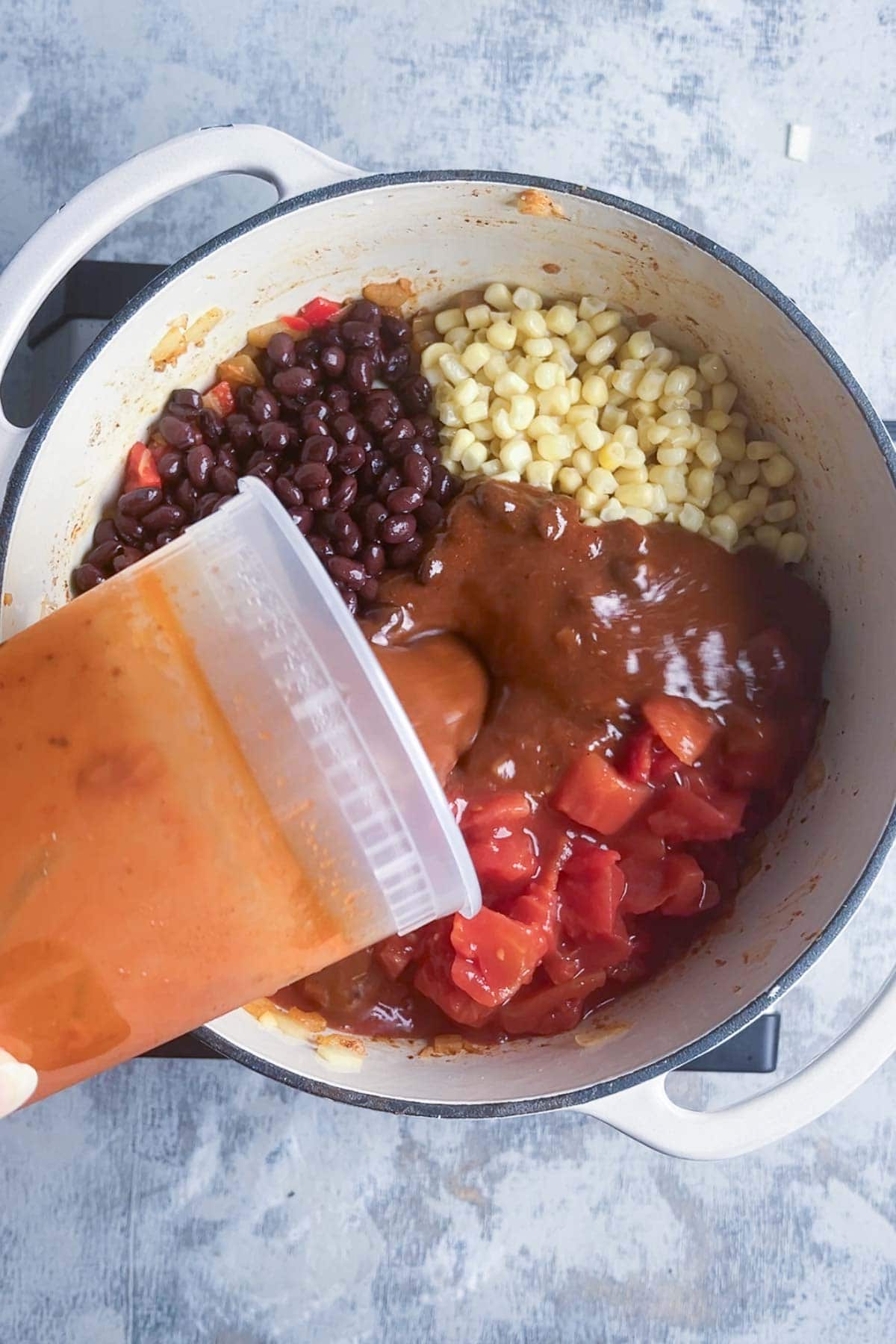 Enchilada sauce is added to a pot filled with black beans, corn, and diced tomatoes.