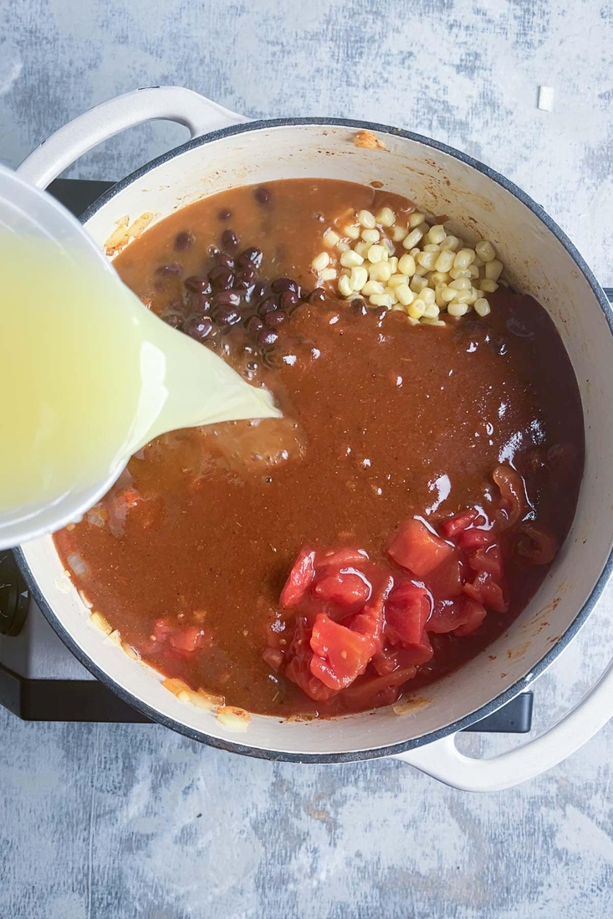 Chicken broth is added to a pot filled with enchilada sauce, black beans, corn, and diced tomatoes.