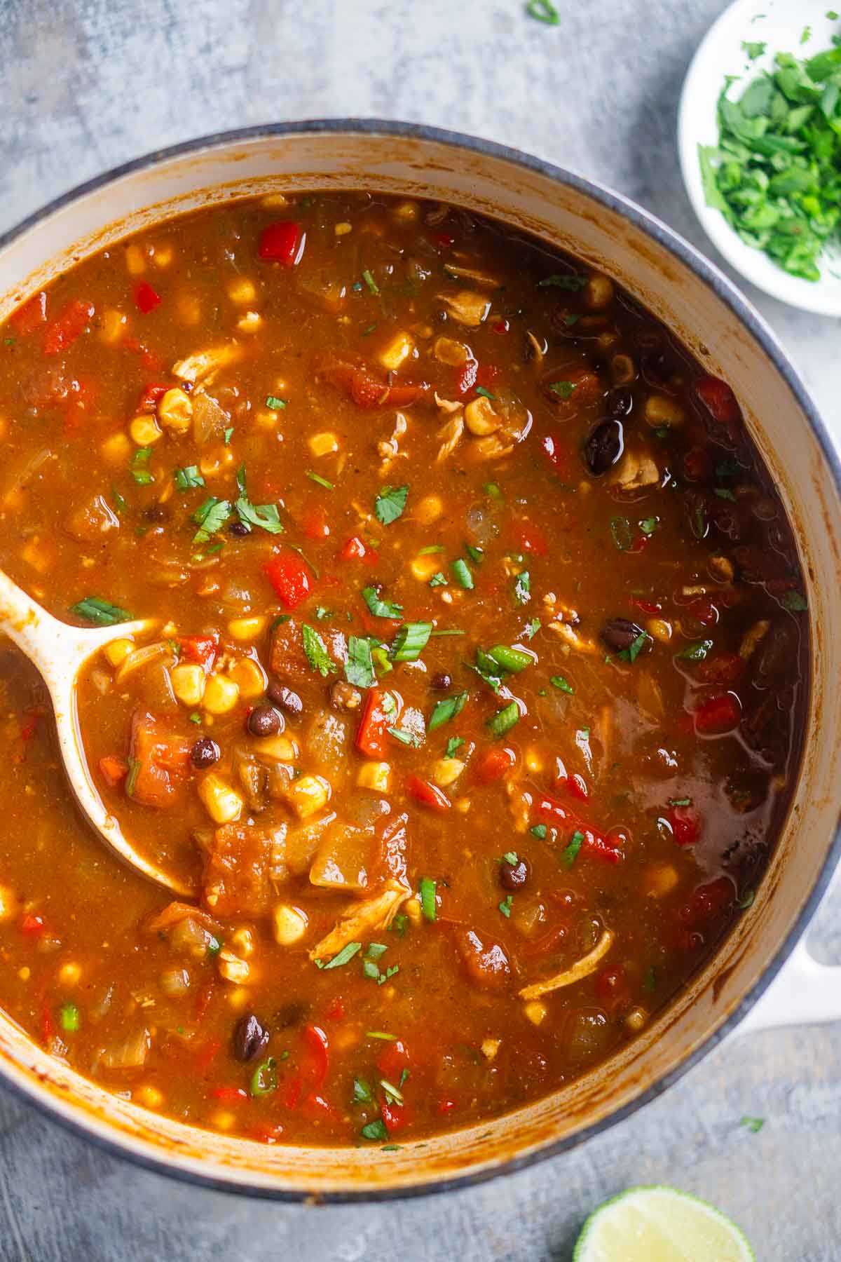 A pot of soup with shredded chicken, black beans, corn, red peppers, saucy red broth, and chopped herbs, with a serving spoon inside.