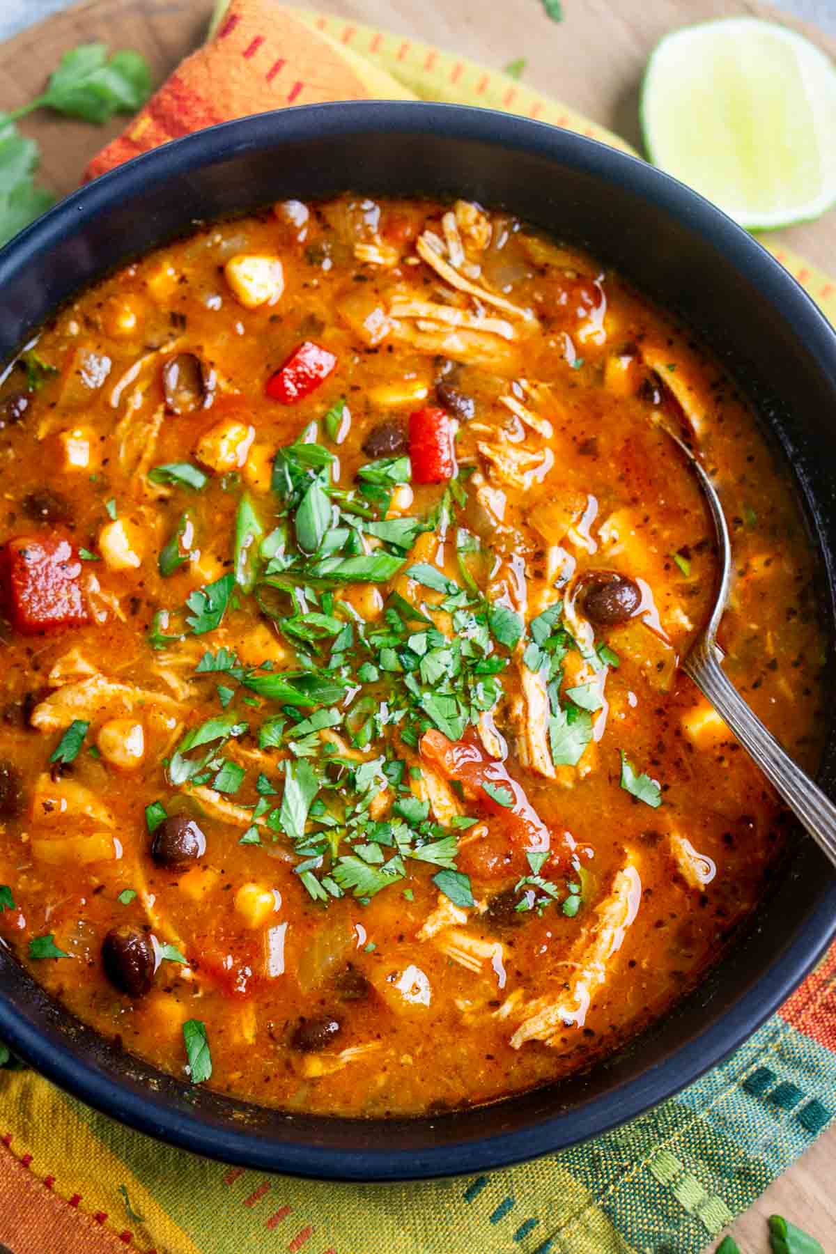 Bowl of chicken tortilla soup topped with chopped cilantro, containing shredded chicken, beans, corn, and red bell peppers, with a spoon inside.
