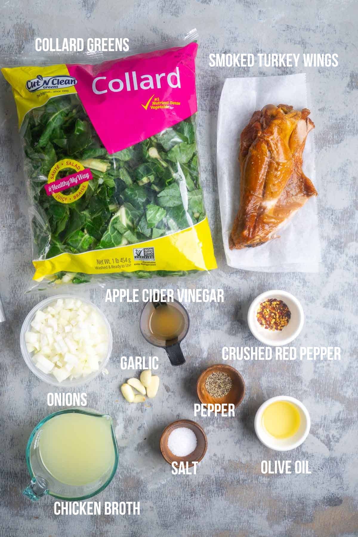 A bag of fresh collard greens, smoked turkey wings, diced onions, garlic cloves, apple cider vinegar, chicken broth, olive oil, salt, pepper, and crushed red pepper are neatly arranged on a gray surface.