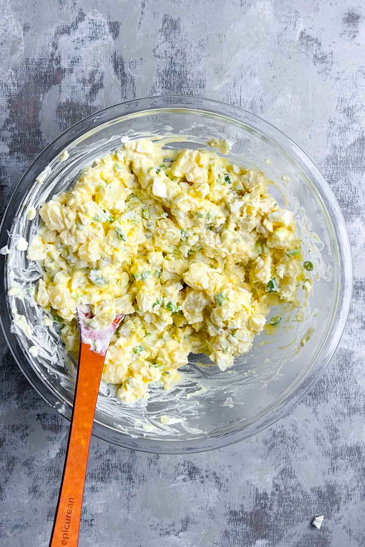 A glass bowl filled with deviled egg potato salad mixed with mayonnaise and herbs, with an orange spatula resting inside the bowl on a gray textured surface.