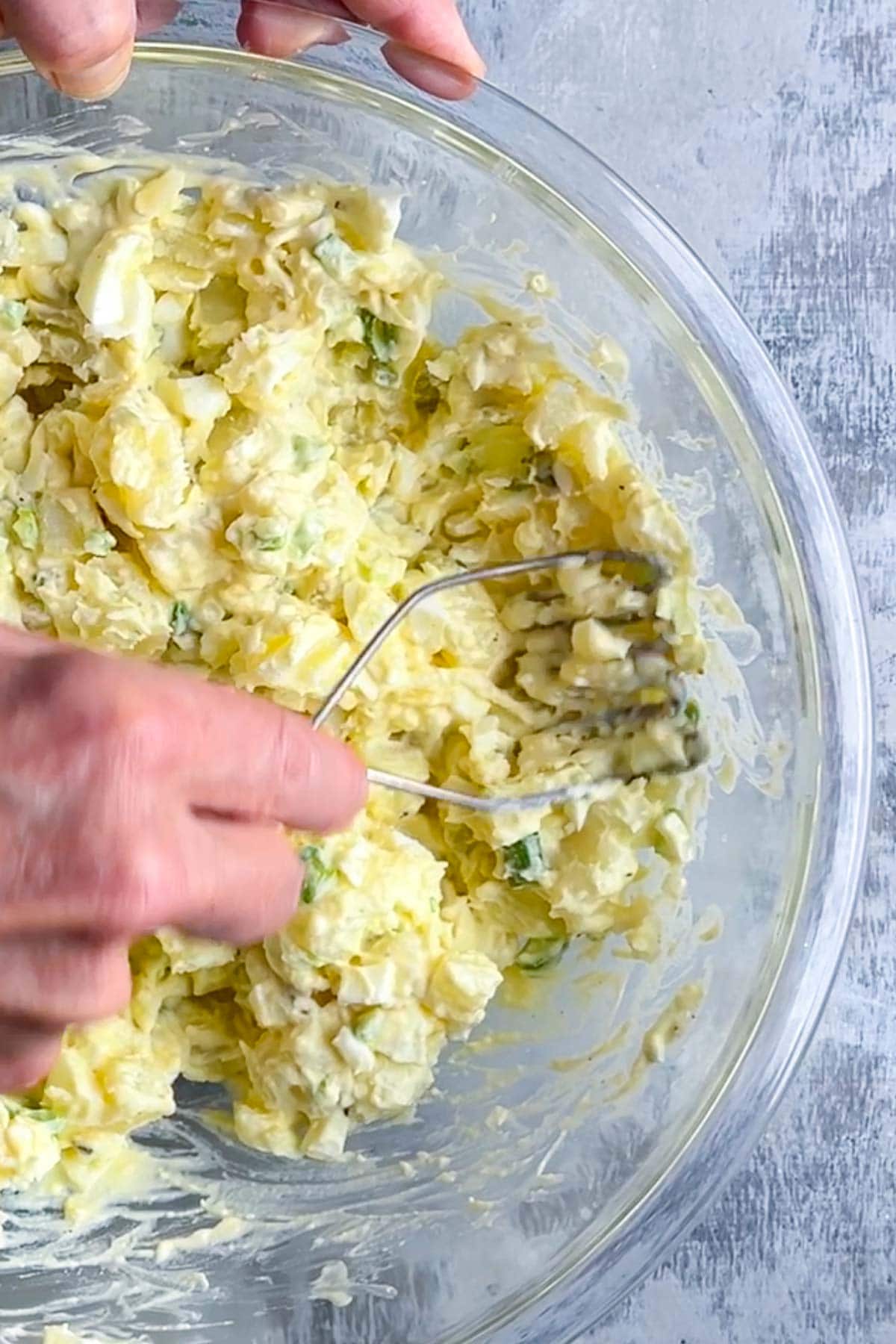 A hand uses a potato masher to mash potato chunks in the deviled egg potato salad into a creamy mixture.