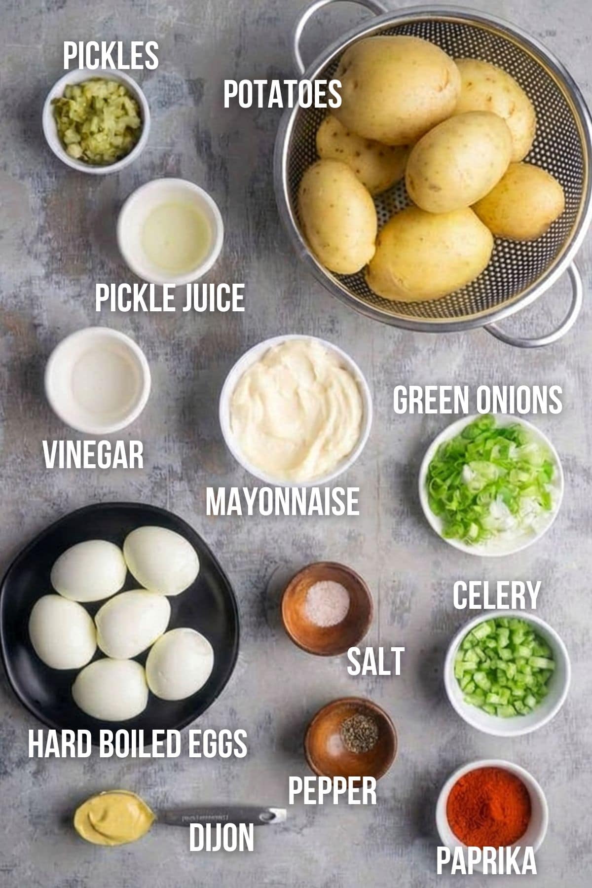 Ingredients for deviled egg potato salad are arranged on a gray surface: potatoes, pickles, pickle juice, vinegar, mayonnaise, green onions, celery, hard boiled eggs, Dijon mustard, salt, pepper, and paprika.