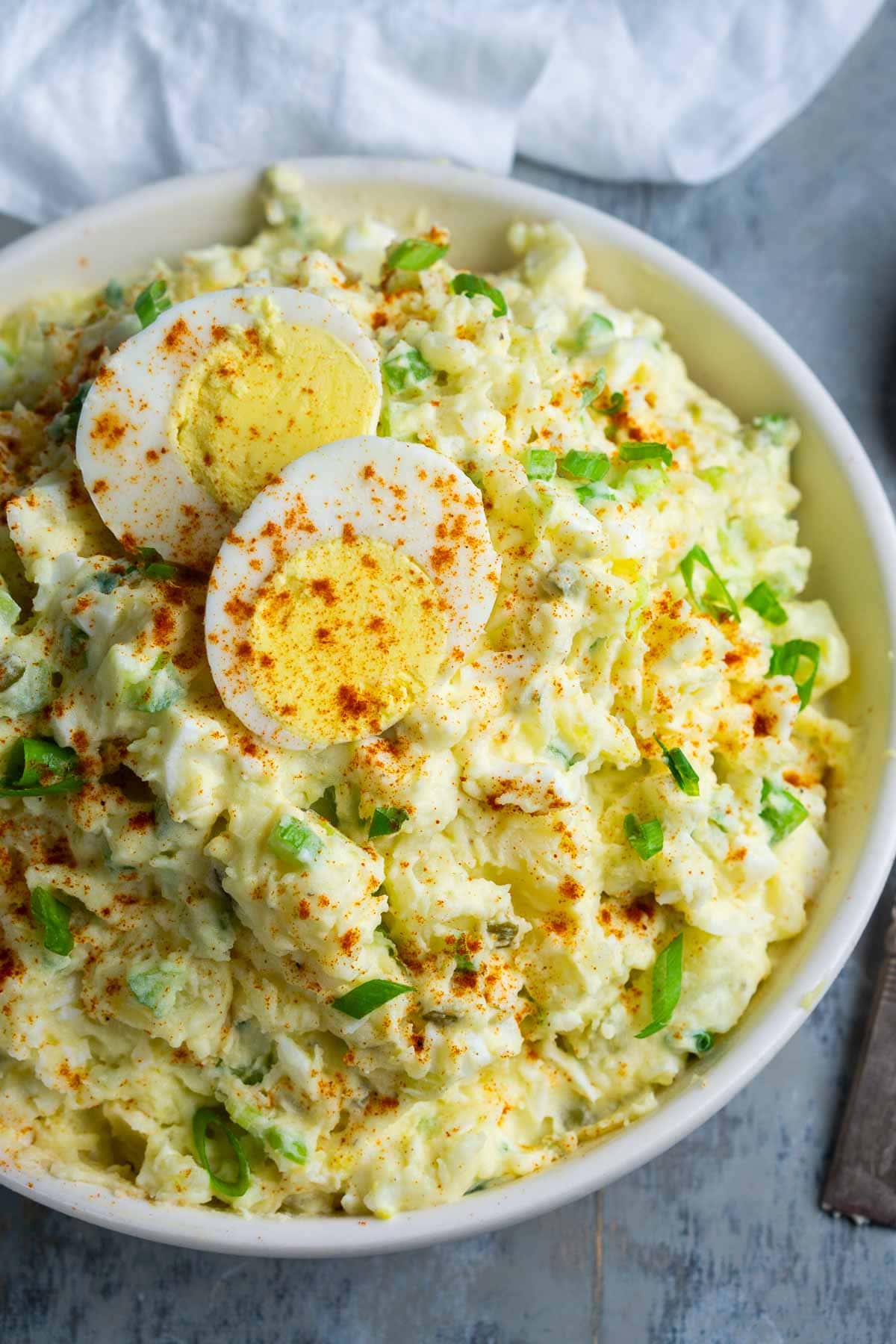 A bowl of deviled egg potato salad topped with sliced boiled eggs, sprinkled with paprika and chopped green onions, with a spoon resting in the bowl.