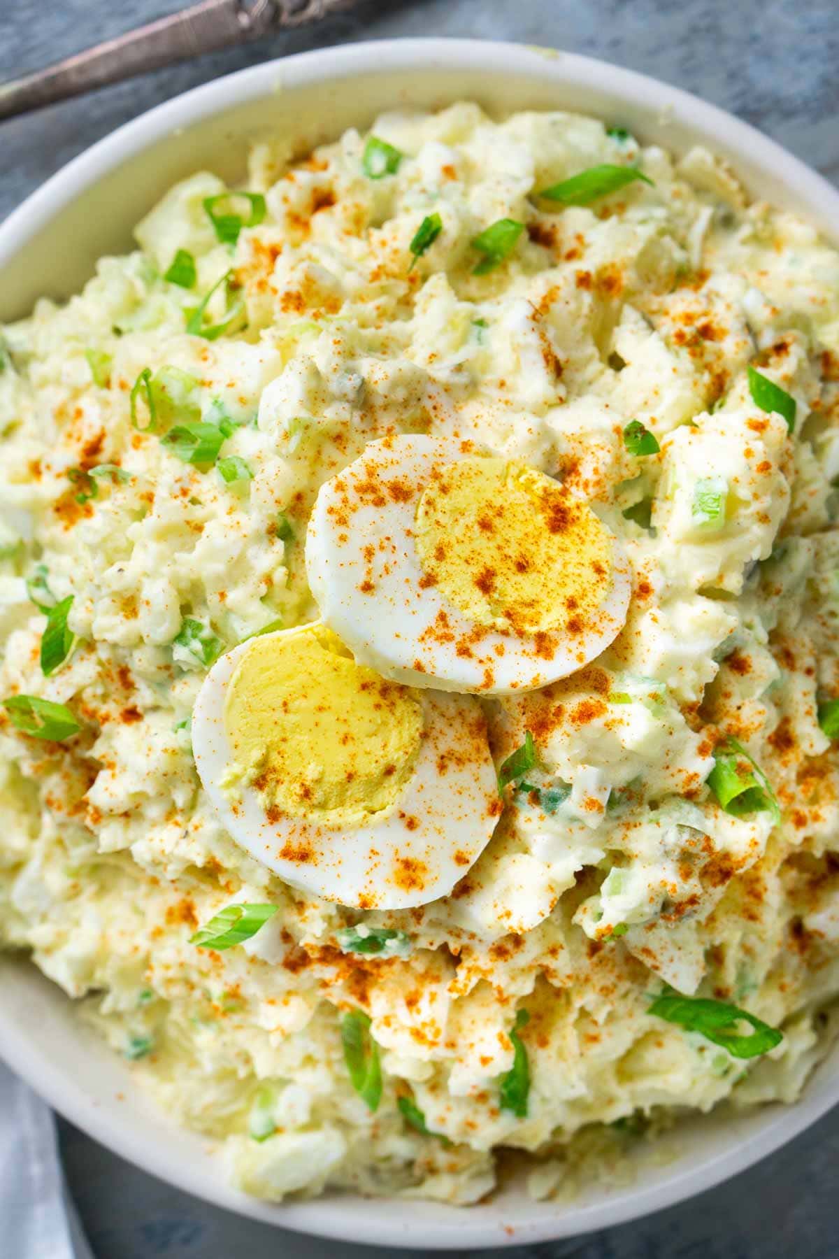 A bowl of deviled egg potato salad topped with sliced boiled eggs, sprinkled with paprika and chopped green onions, with a spoon resting in the bowl.