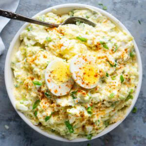 A bowl of deviled egg potato salad topped with sliced boiled eggs, sprinkled with paprika and chopped green onions, with a spoon resting in the bowl.