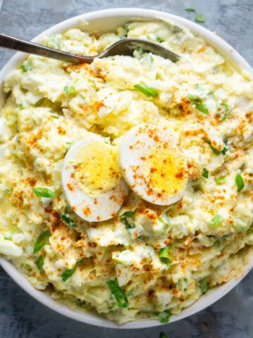 A bowl of deviled egg potato salad topped with sliced boiled eggs, sprinkled with paprika and chopped green onions, with a spoon resting in the bowl.