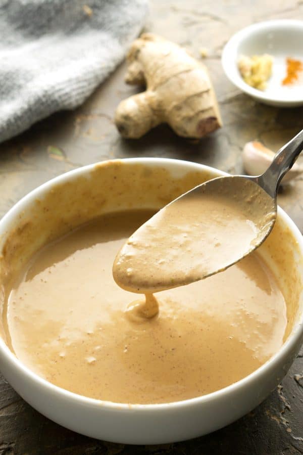 Spoonful of Thai peanut sauce over white bowl on table surface with fresh ginger, garlic, and dry spices in small white bowl.
