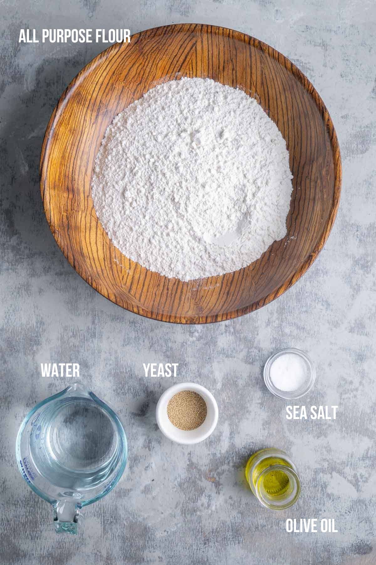 A wooden bowl with flour, ready for focaccia bread, is surrounded by small containers of water, yeast, sea salt, and olive oil on a gray surface. Labels identify each ingredient.