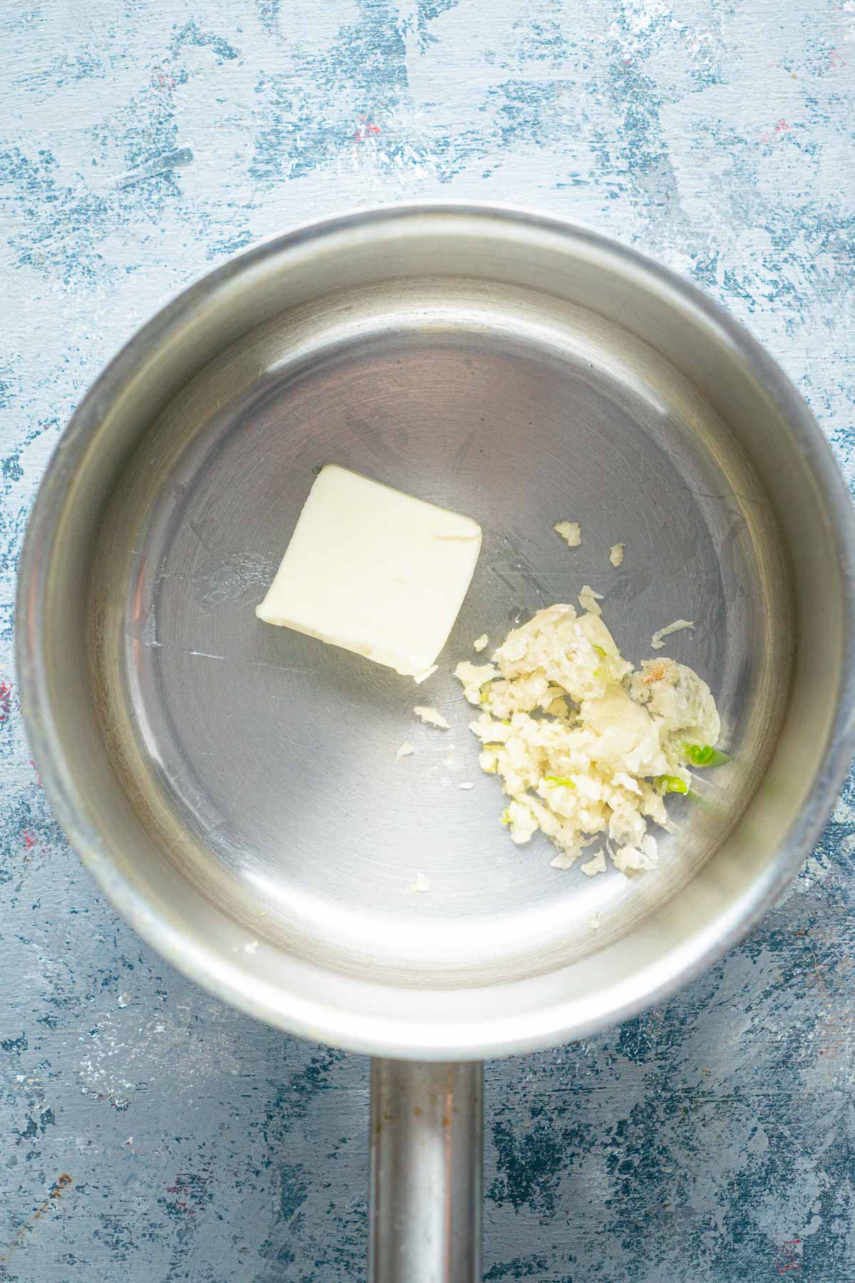 A saucepan with a pat of butter and a small pile of minced garlic on a light textured surface.