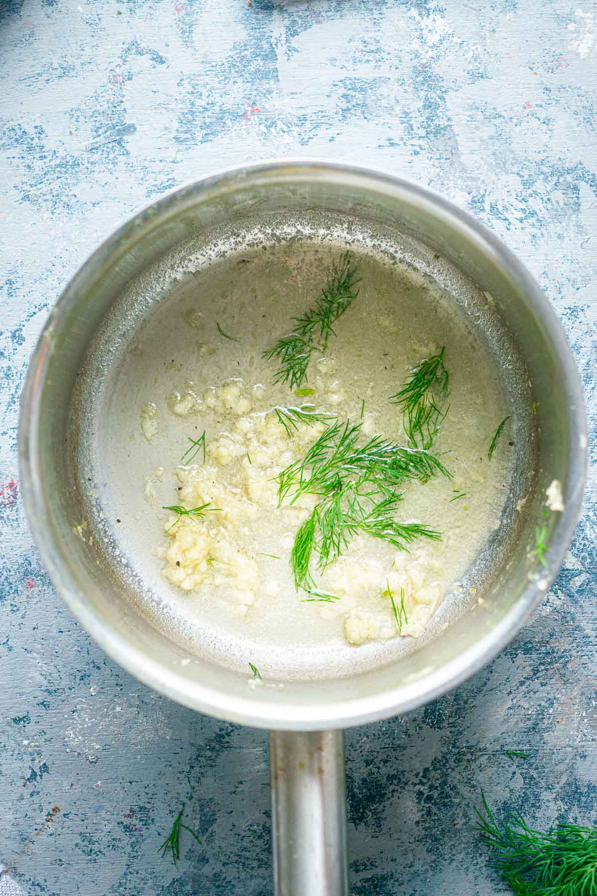 A saucepan with melted butter, minced garlic, and fresh dill on a textured light blue surface.