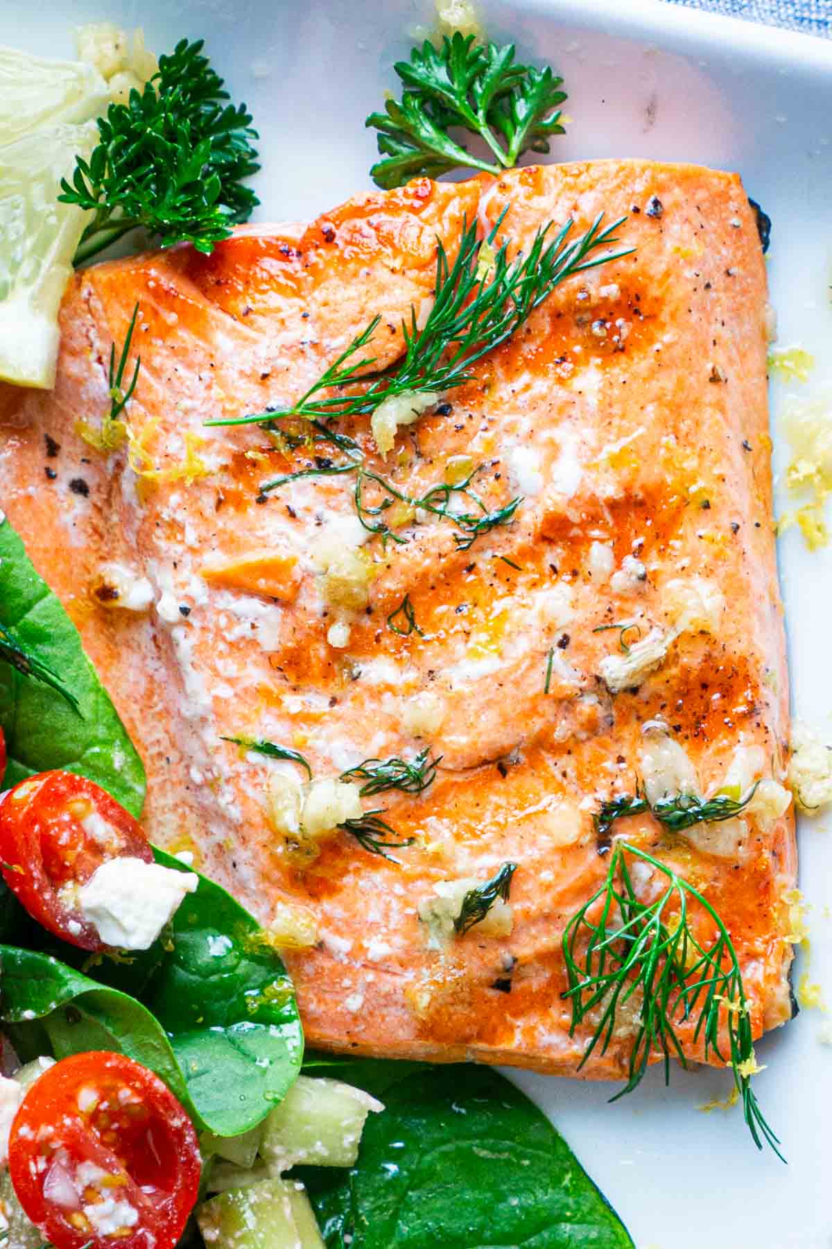 A buttery grilled salmon fillet garnished with fresh dill, parsley, and lemon wedge, served next to a salad on a white plate.