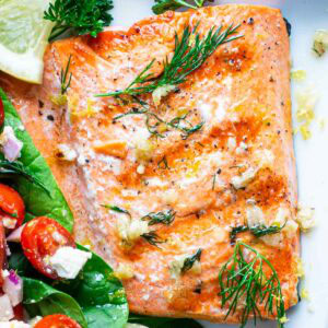 A buttery grilled salmon fillet garnished with fresh dill, parsley, and lemon wedge, served next to a salad on a white plate.
