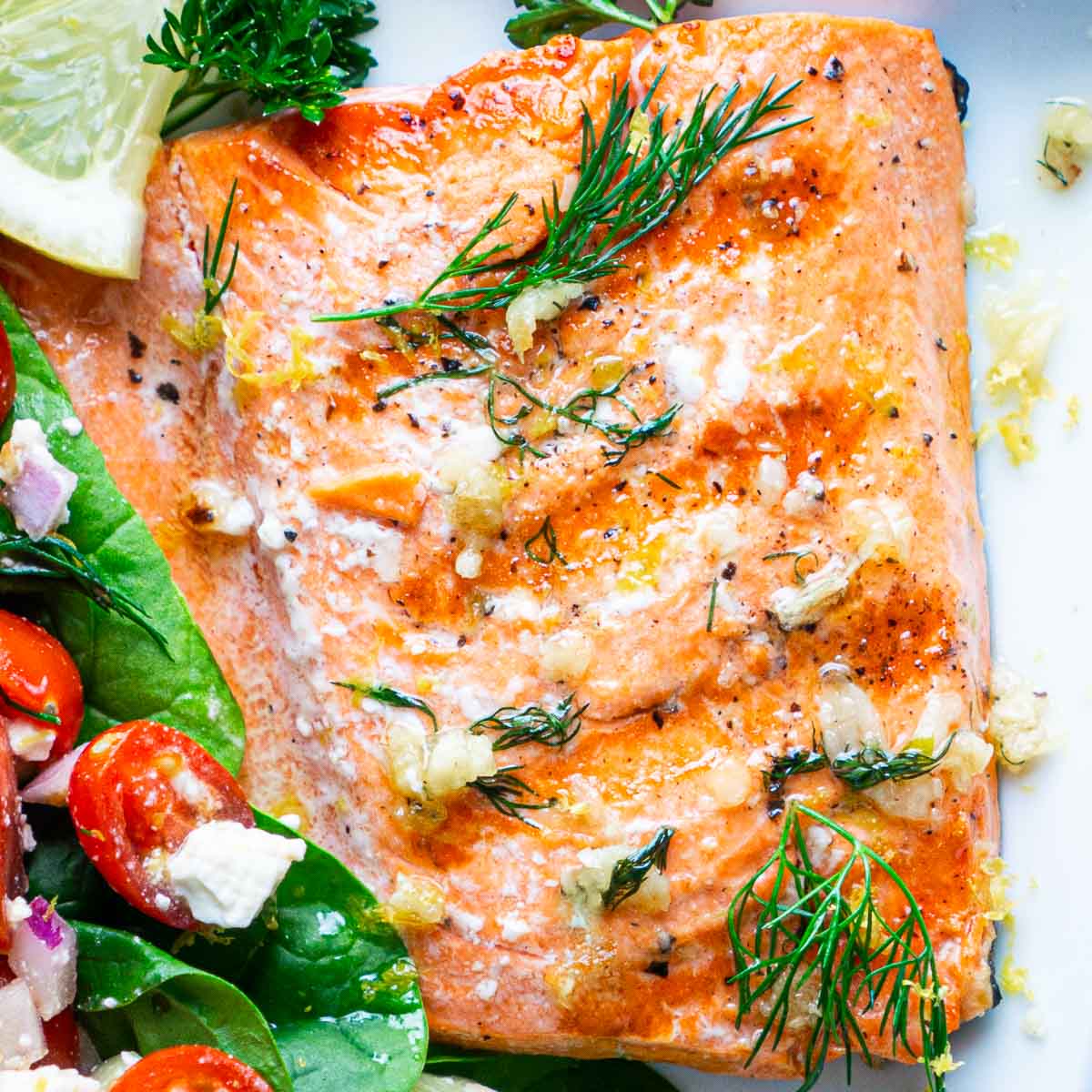 A buttery grilled salmon fillet garnished with fresh dill, parsley, and lemon wedge, served next to a salad on a white plate.