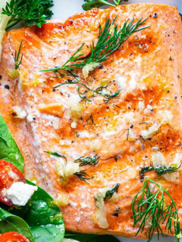 A buttery grilled salmon fillet garnished with fresh dill, parsley, and lemon wedge, served next to a salad on a white plate.