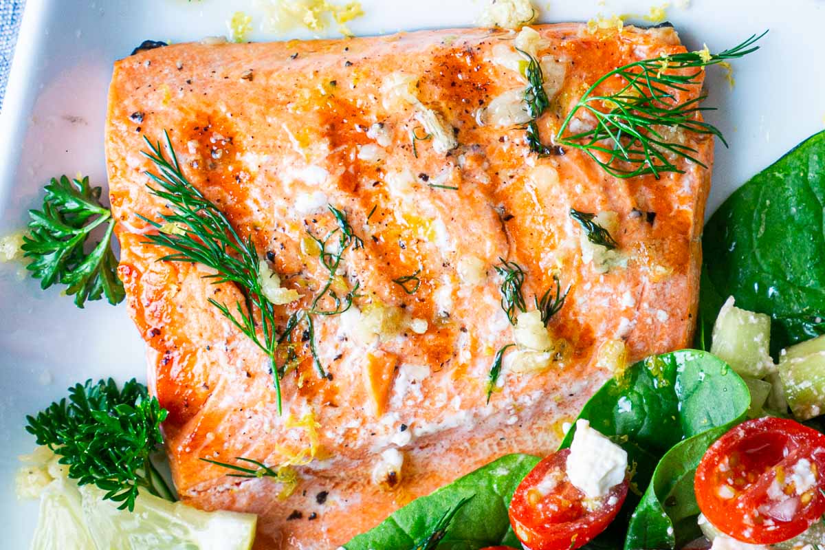 A buttery grilled salmon fillet garnished with fresh dill, parsley, and lemon wedge, served next to a salad on a white plate.