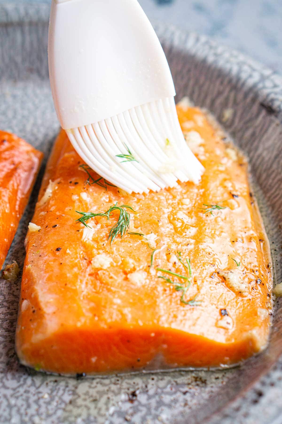A close-up of a raw salmon fillet being brushed with dill garlic butter, on a gray plate.