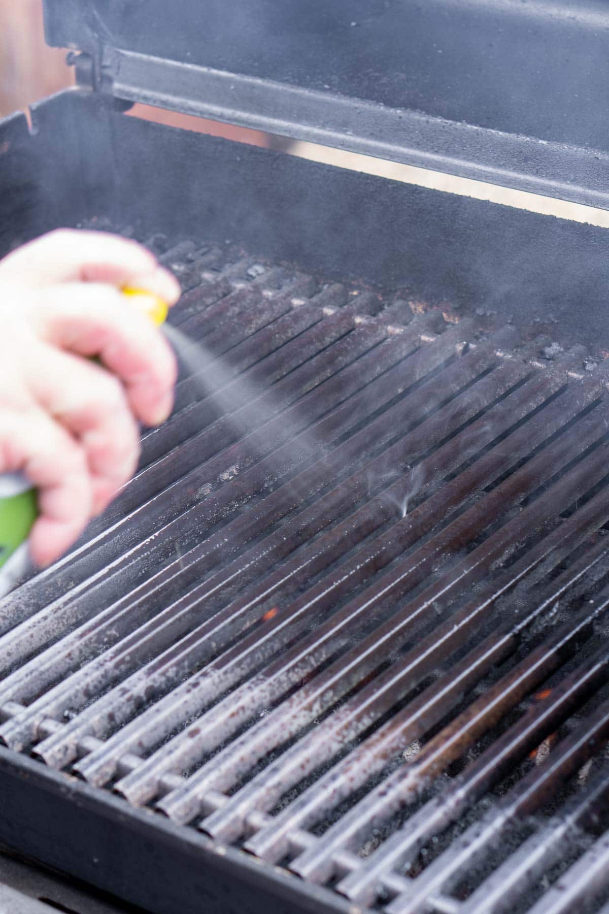 A person sprays oil onto the grates of an outdoor grill, which is open and emitting some smoke.