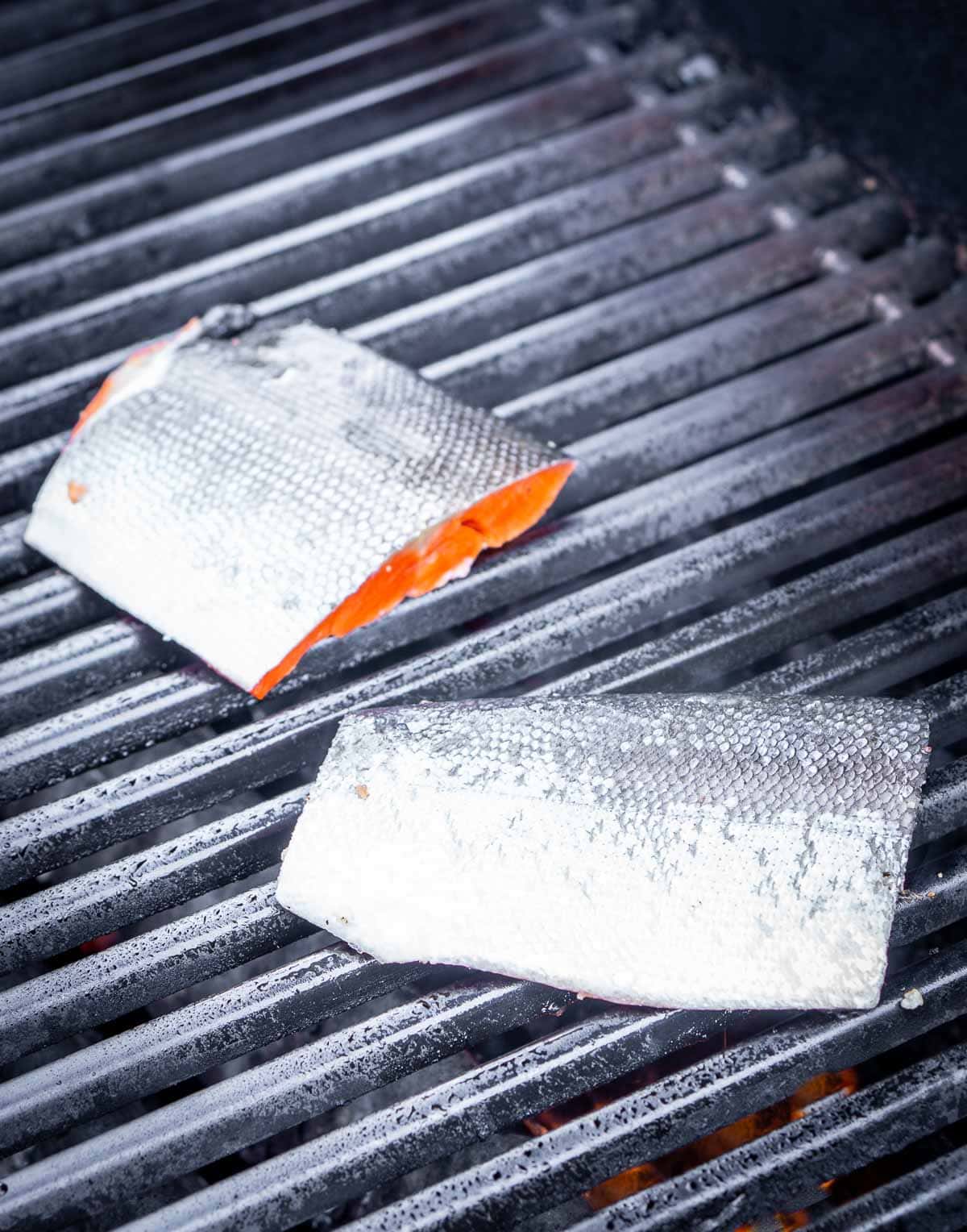 Two pieces of grilled salmon fillet with skin on are cooking on a metal grill.