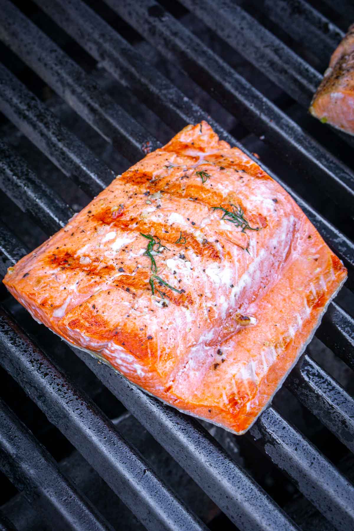 A piece of grilled salmon fillet, perfectly seasoned, sizzles on a black grill grate as it develops beautiful charred grill marks.