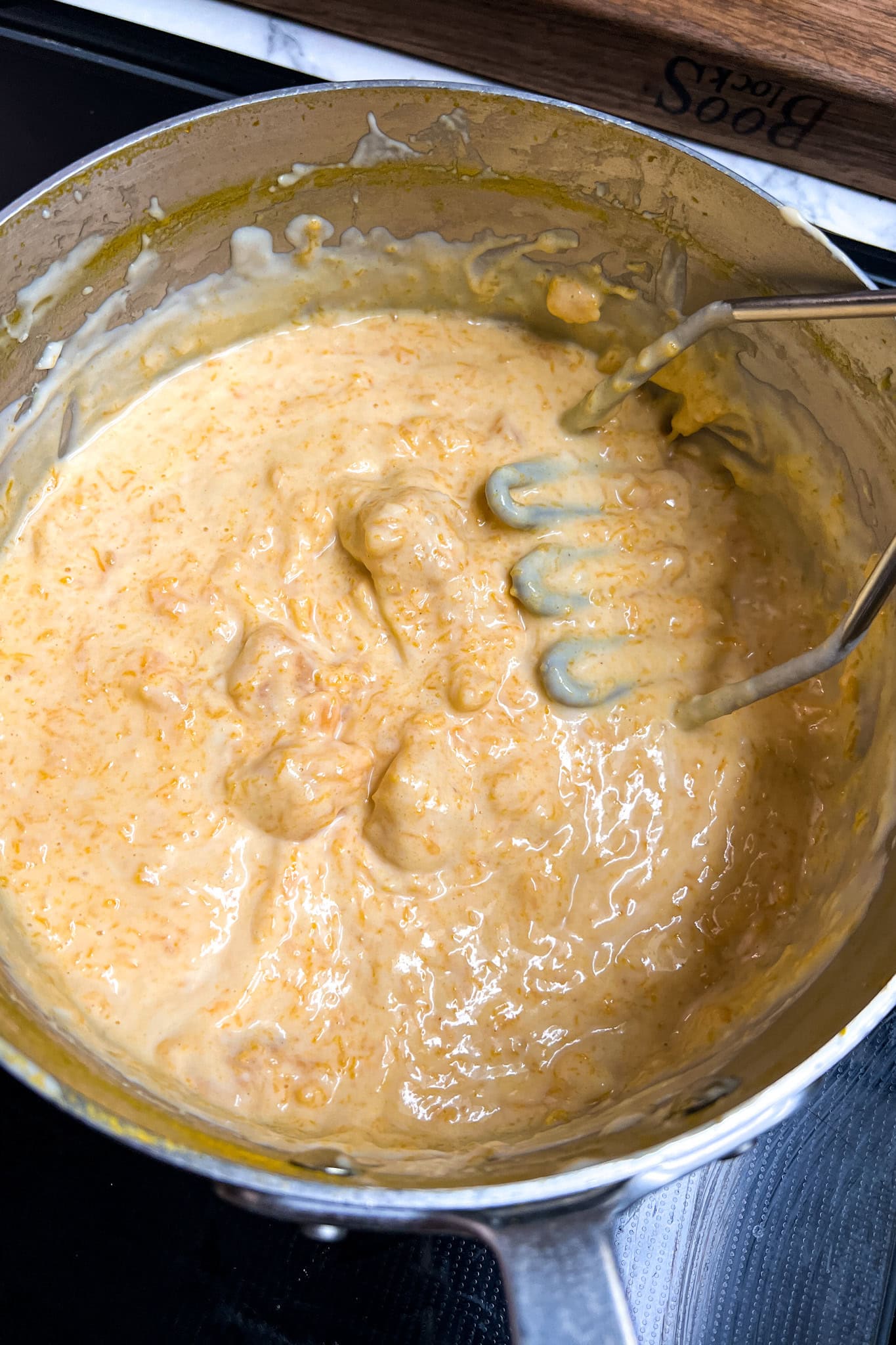 A pot of thick, creamy cheese mixture in a saucepan with a vegetable masher resting inside.