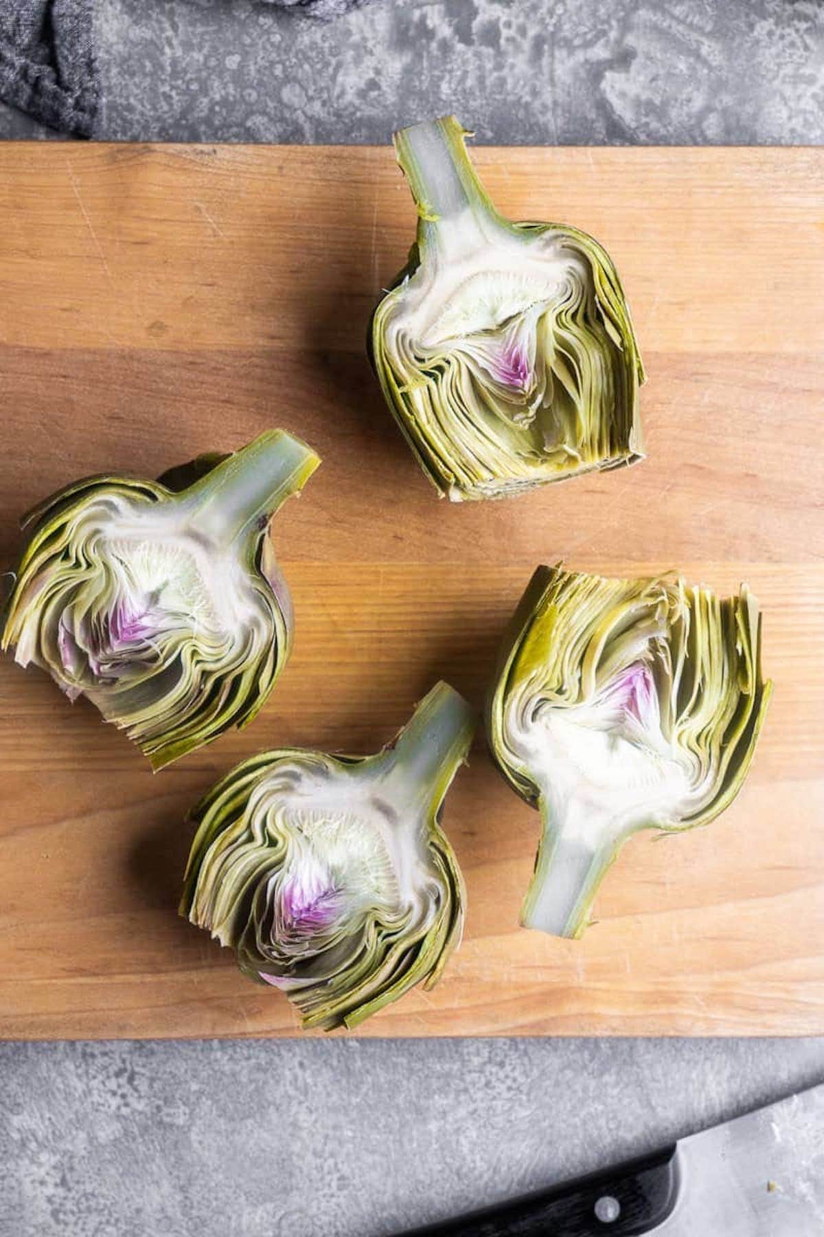 Four steamed artichokes cut in half are arranged on a wooden cutting board, displaying their inner layers and purple centers. A knife is partially visible at the bottom right.
