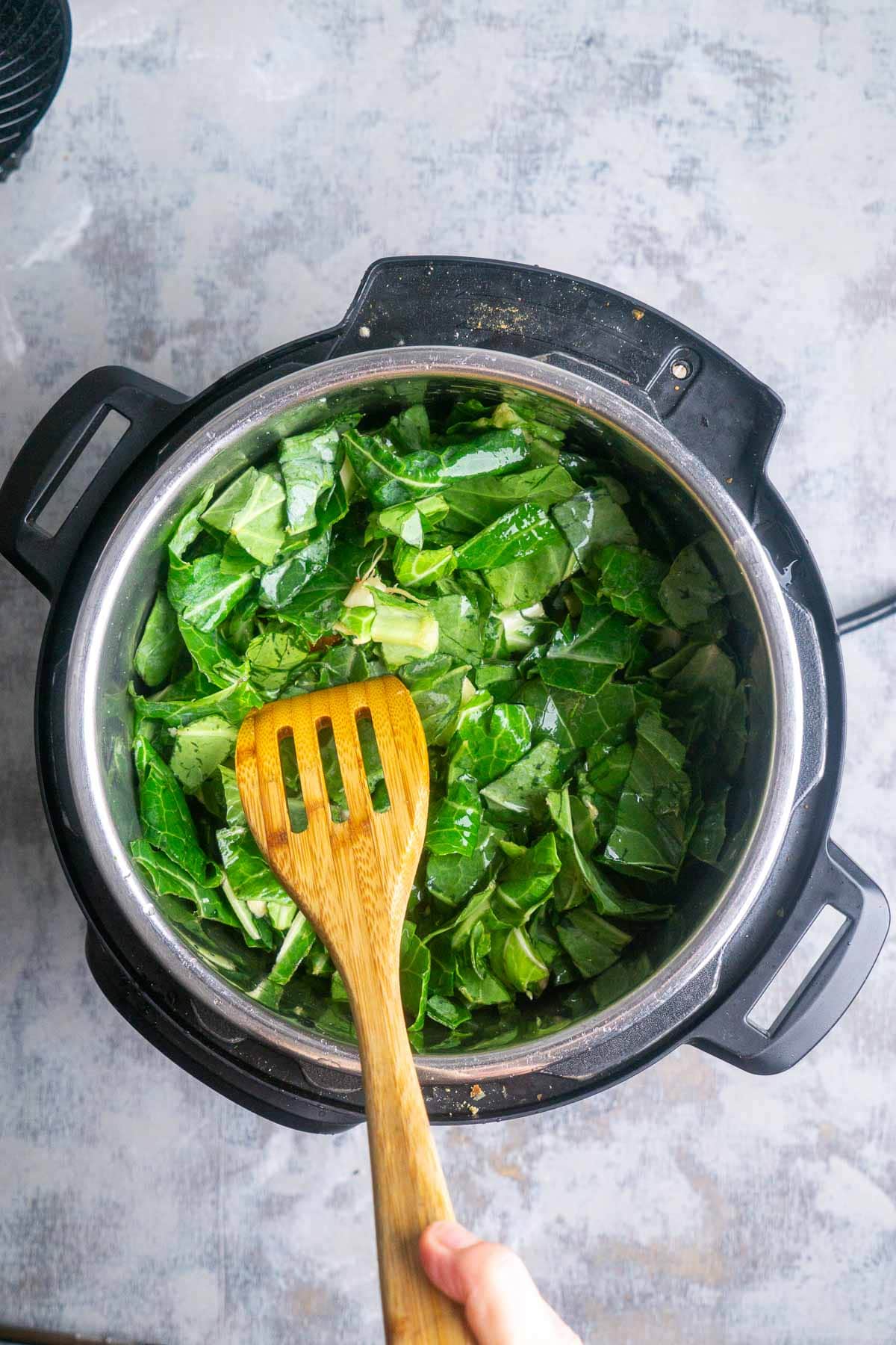 A person's hand holding a wooden utensil pushes down chopped collard greens inside an Instant Pot on a light-colored surface.