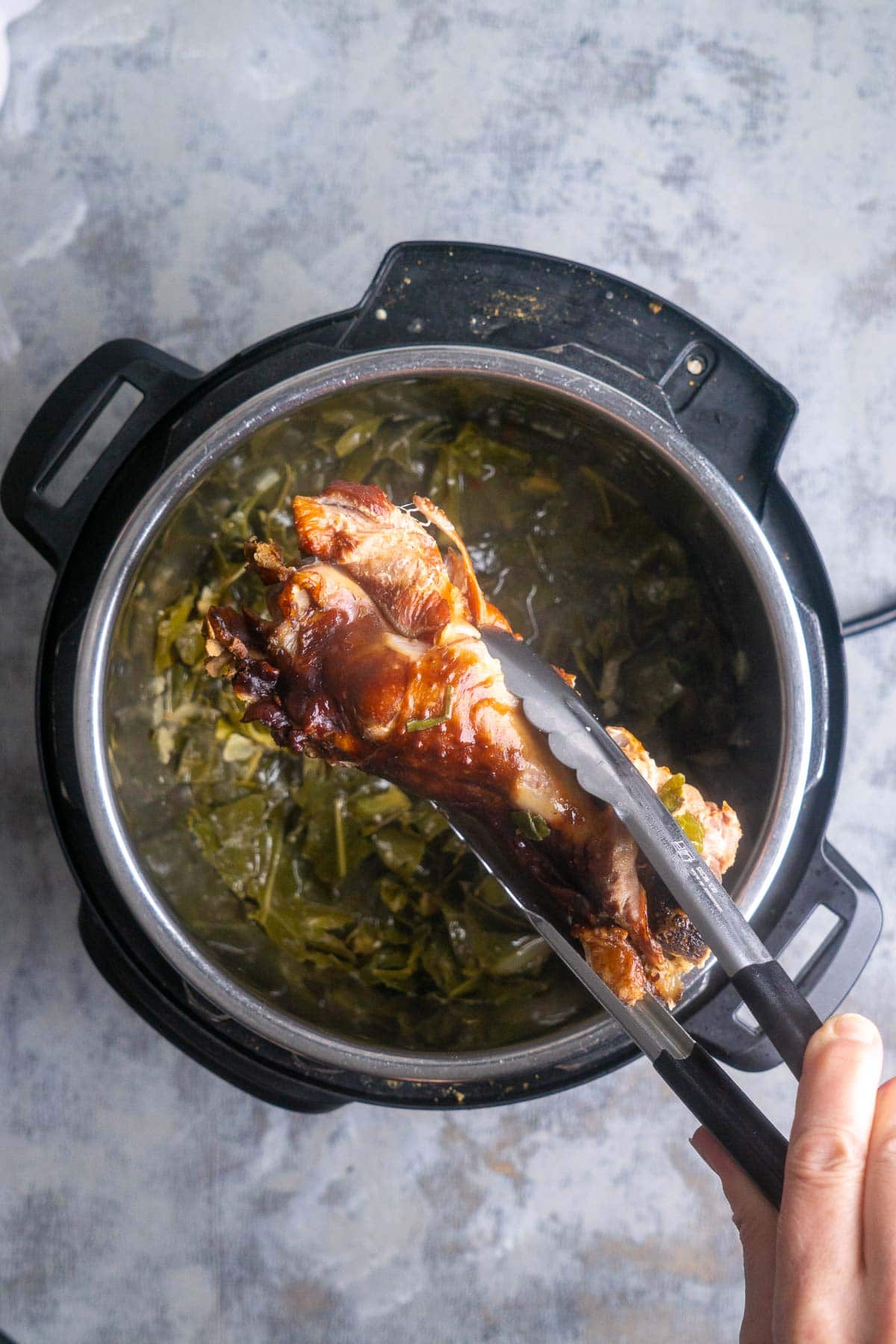 A hand uses tongs to remove a cooked turkey leg from an Instant Pot filled with collard greens and broth.