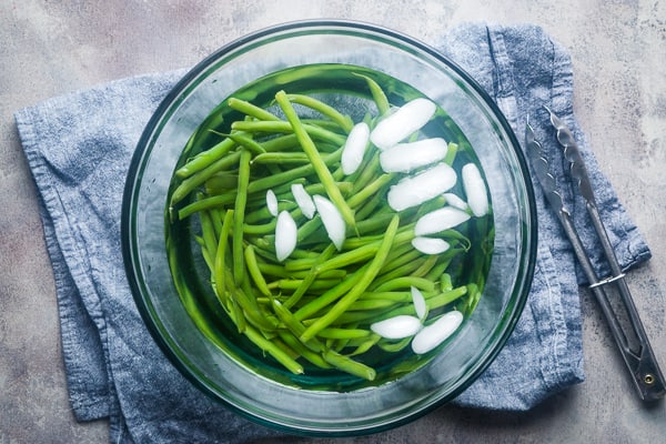 green beans and ice water in glass bowl next to tongs and blue linen