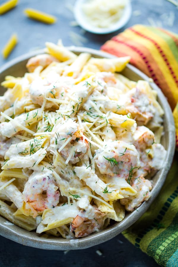 creamy shrimp pasta in bowl with multicolored napkin and penne shrimp pieces and white bowl of parmesan cheese on blue background