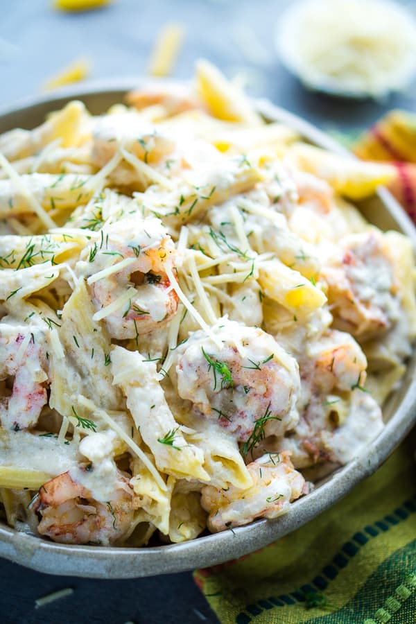 creamy shrimp pasta in bowl with multicolored napkin and penne shrimp pieces and white bowl of parmesan cheese on blue background.