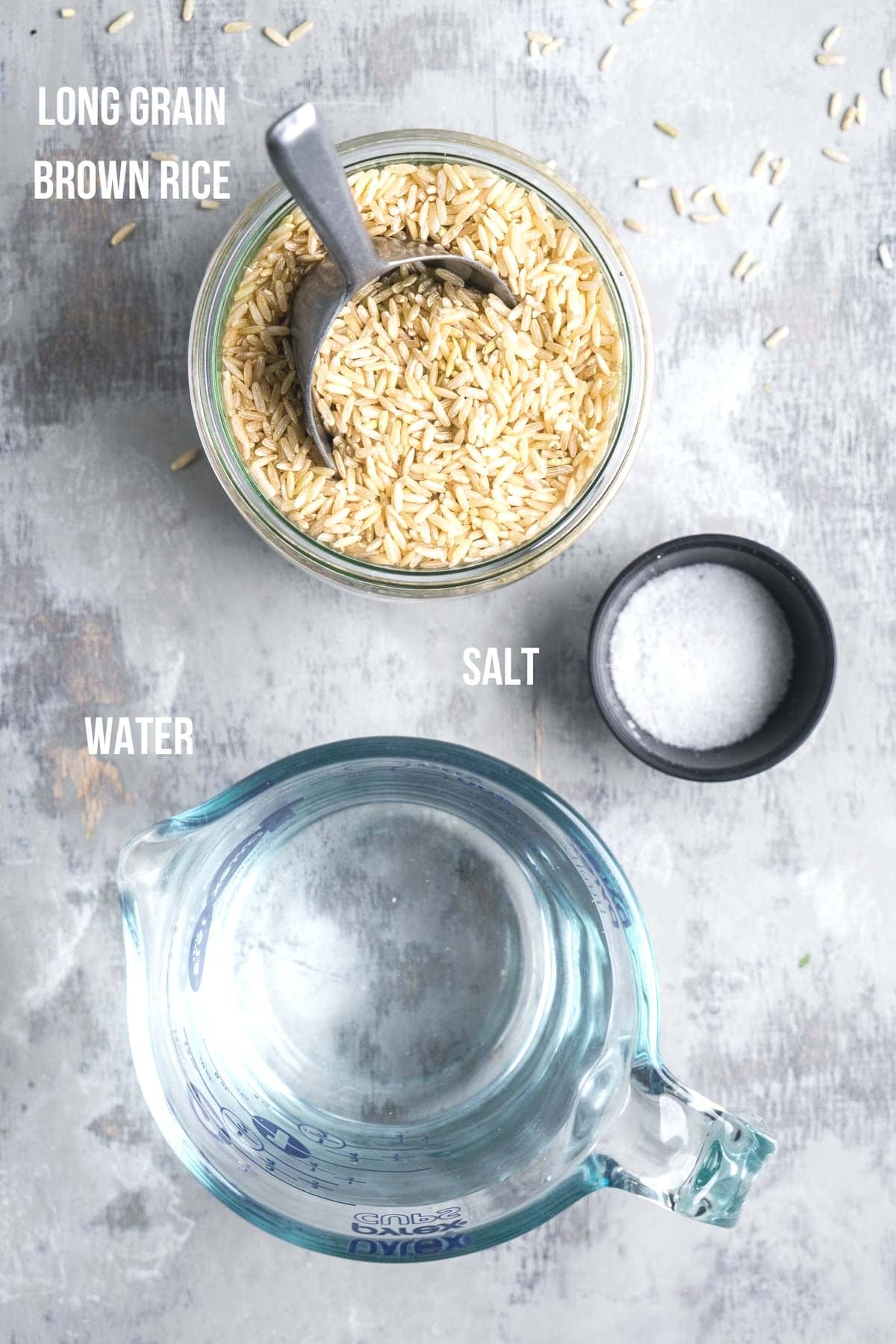Top-down view of a jar of long grain brown rice with a scoop, a measuring cup of water, and a small bowl of salt on a gray surface.