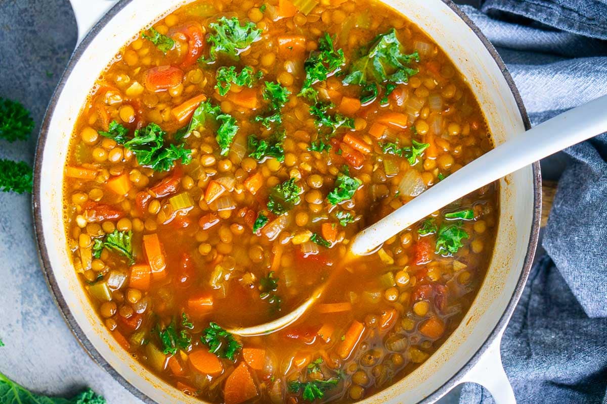 A white pot of Italian lentil soup with lentils, carrots, tomatoes, and kale, with a white ladle stirring the ingredients.