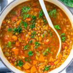 A white pot of Italian lentil soup with lentils, carrots, tomatoes, and kale, with a white ladle stirring the ingredients.