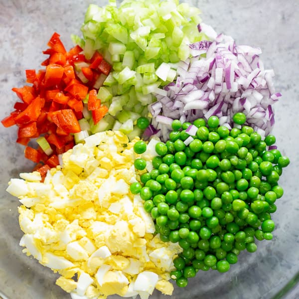 macaroni salad ingredients in glass bowl (diced red bell pepper, celery, red onion, hard-boiled eggs, and green peas)