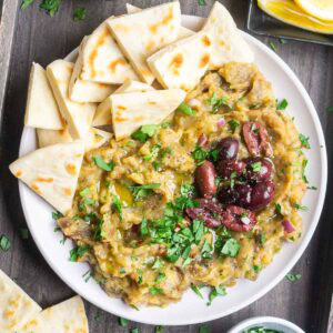 A plate of Greek eggplant dip on white plate garnished with parsley and olives, surrounded by sliced pita bread on a wooden surface.