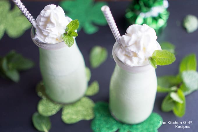 shamrock shake in glass bottles with straw, whipped cream, and mint leaf garnish next to St Patrick's Day decorations
