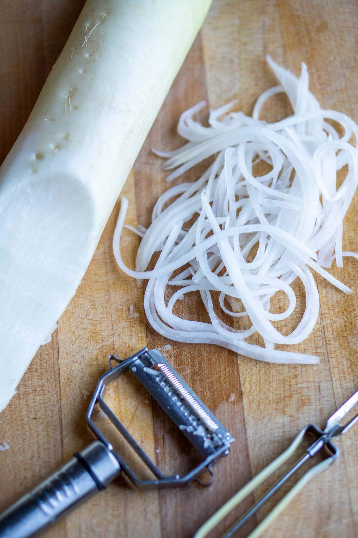 Daikon radish partially peeled and julienned on a wooden cutting board with a julienne peeler and a regular peeler beside it.
