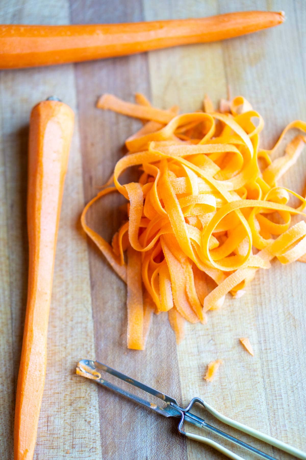 Two peeled carrots, a pile of carrot ribbons, and a vegetable peeler on a wooden cutting board.
