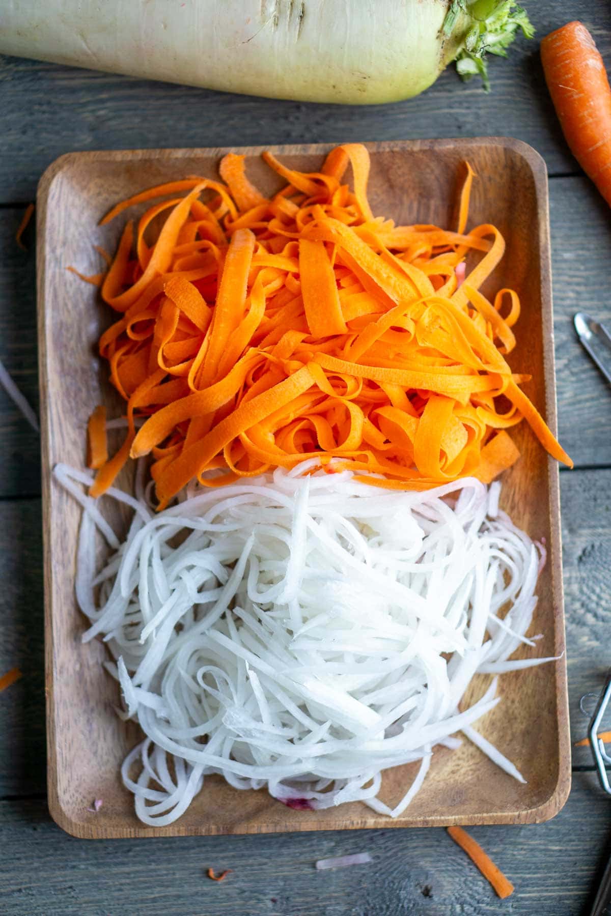 A wooden tray holding a pile of shredded white daikon and a pile of orange carrot ribbons, with a daikon and carrot nearby on a wooden surface.